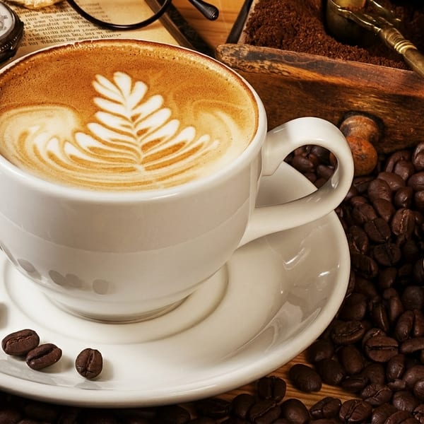 A cup of latte with leaf art surrounded by coffee beans, an open book, and a pocket watch.